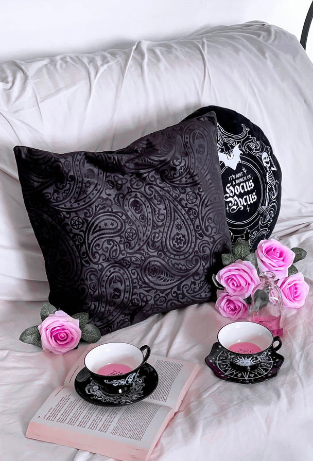 A black pillow with a paisley design laying on a bed with pink roses, a tea cup sitting on a saucer next to a book.