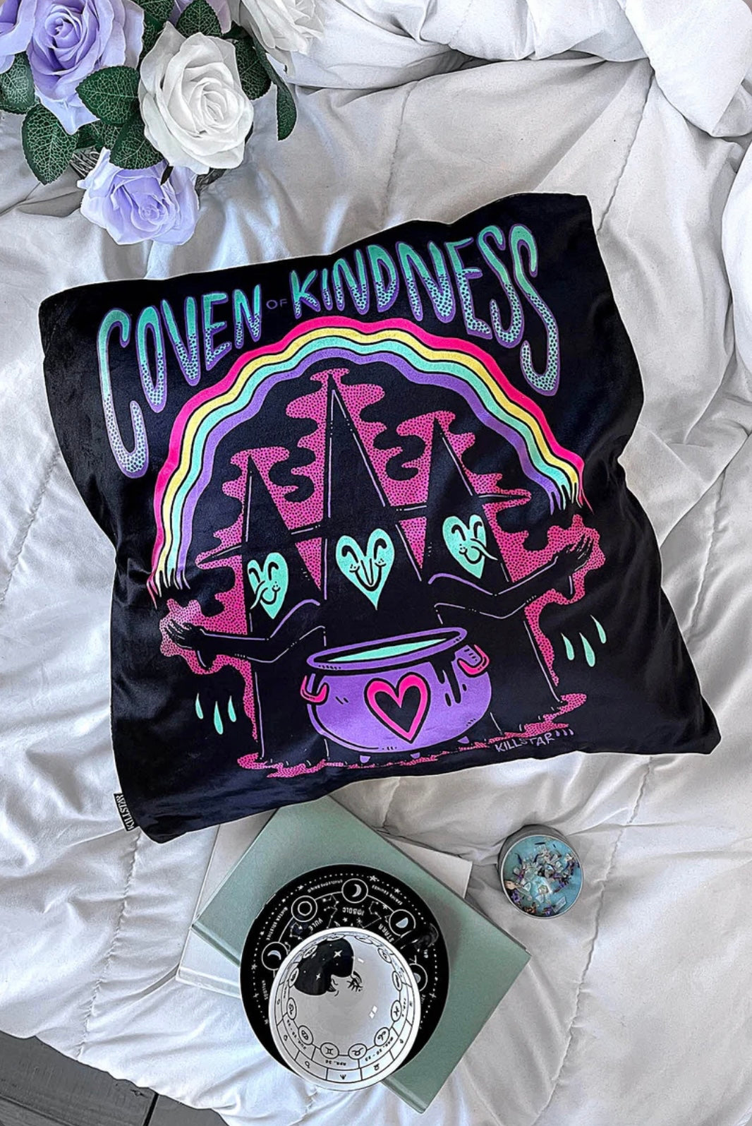 A black pillow cushion with rainbow colors featuring a witch, purple pot and Coven of Kindness text in aqua and purple.