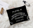 A gothic print makeup bag with the words “ ouija yes no goodbye “ written on it laying on a white fur prop rug.