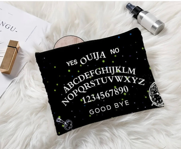 A gothic print makeup bag with the words “ ouija yes no goodbye “ written on it laying on a white fur prop rug.