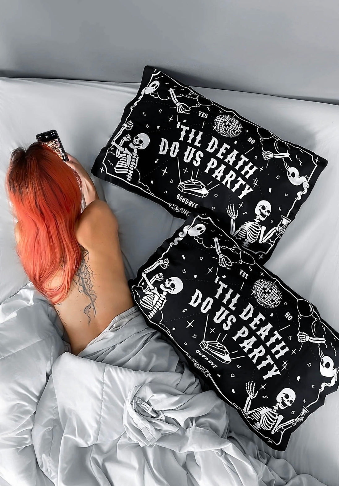 A woman relaxing in bed looking at her cellphone with two black pillows with white text and skeletons laying next to her.