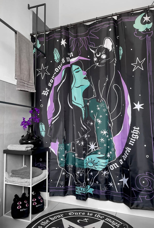 A black shower curtain with white text, a woman, cat, moon and stars hanging on a shower rack in a bathroom.